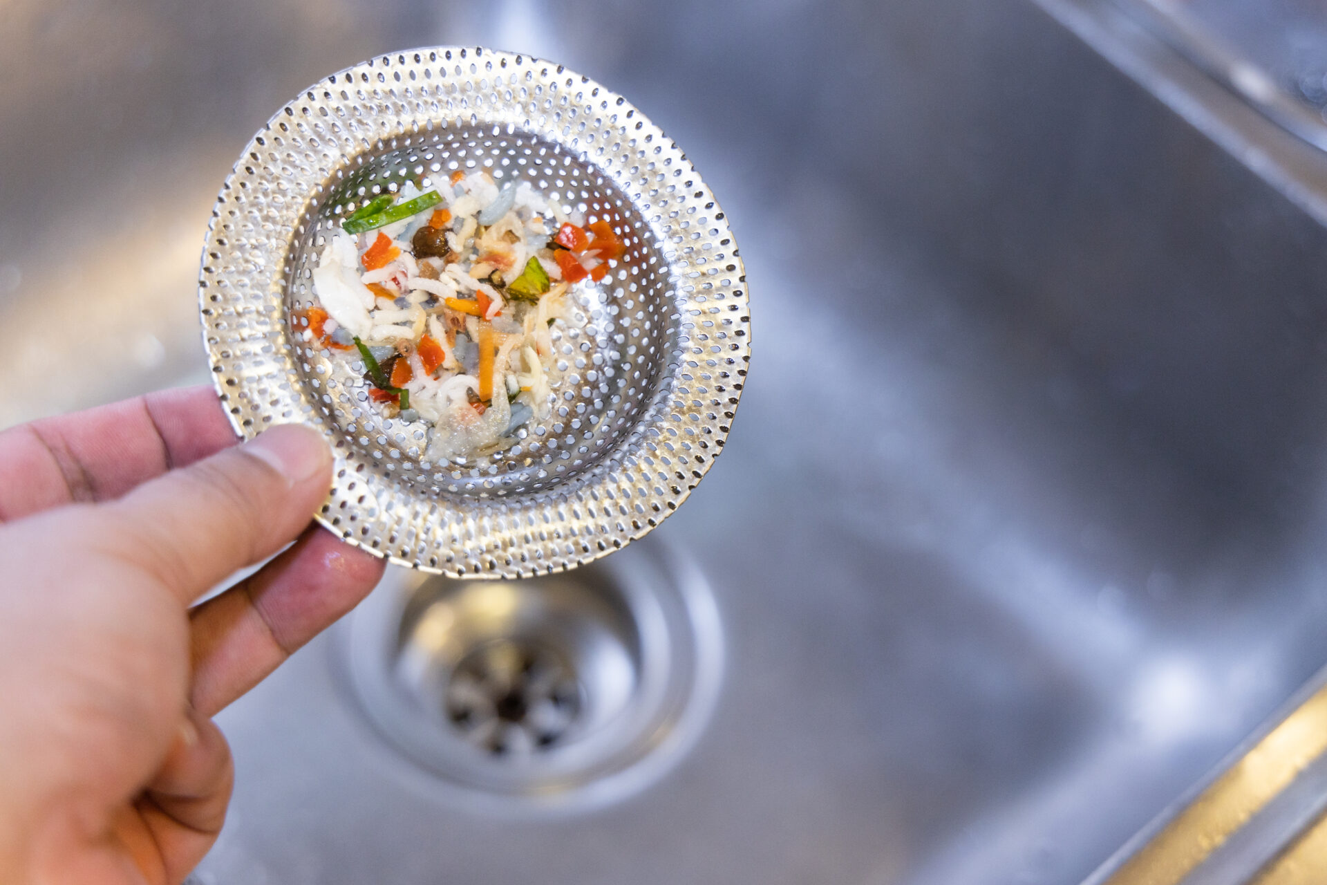 Hand holding kitchen sink waste filter with trapped food waste, against sink background