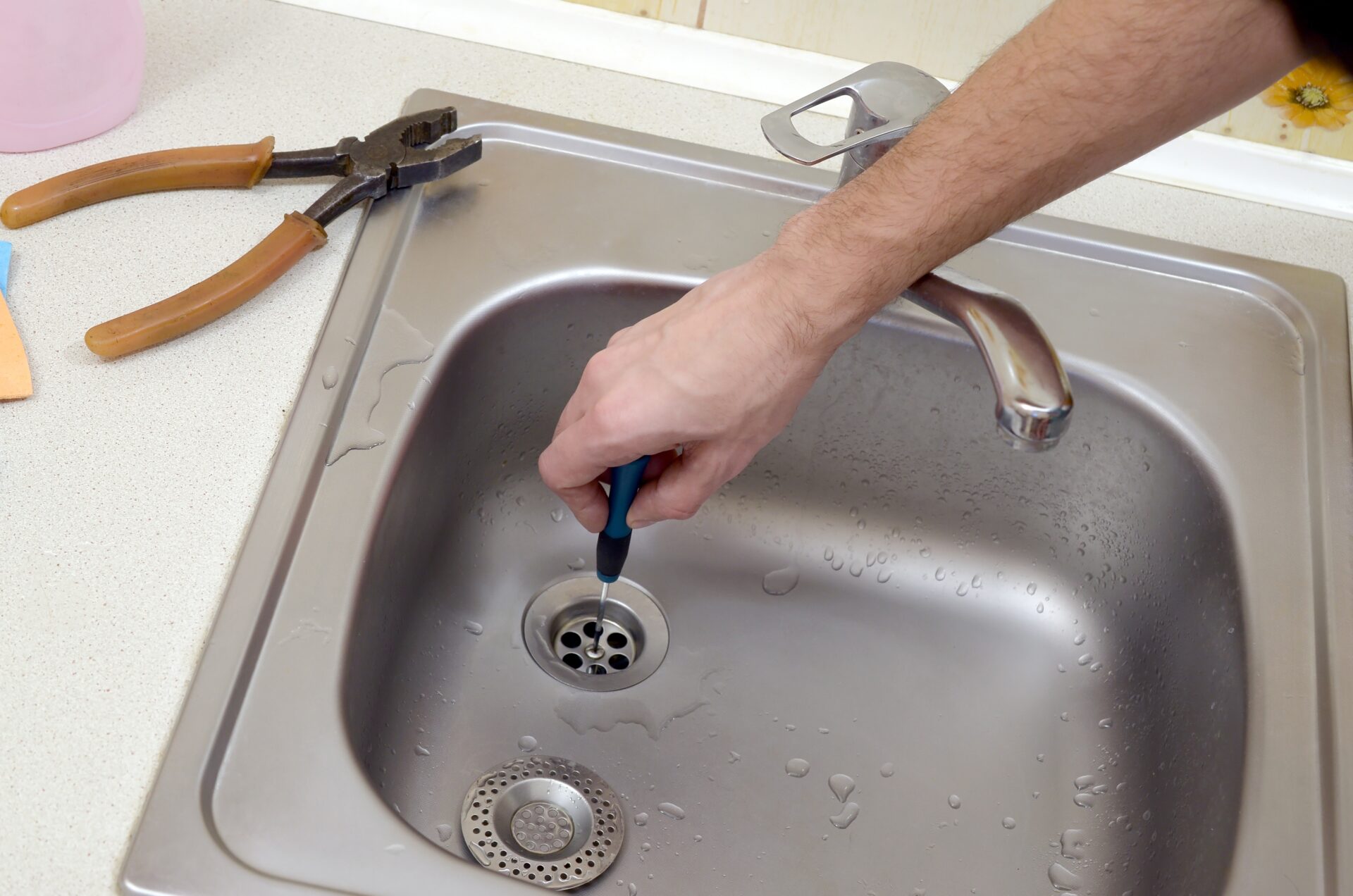 Close-up of handsome plumber repairing drain of kitchen sink. Photo of metalic sink and handyman with screwdriver